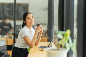 Beautiful young asian business woman drinking coffee and using laptop computer while working in office. photo
