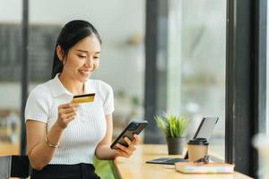 smiling woman paying online, using laptop, holding plastic credit card, sitting on coffee shop, Asian female shopping, making secure internet payment, browsing banking service. photo