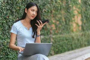 Portrait of beautiful Asian woman sitting outdoors during summer, using smart wireless technology computer laptop and smartphone, relaxing coffee break at cafe restaurant. photo