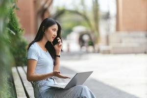 Portrait of beautiful Asian woman sitting outdoors during summer, using smart wireless technology computer laptop and smartphone, relaxing coffee break at cafe restaurant. photo