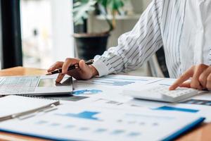 cerca de la mano del empresario o contador mano sosteniendo la pluma trabajando en la calculadora para calcular datos comerciales, documentos contables y computadora portátil en la oficina, concepto de negocio. foto