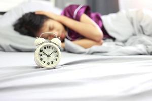 Wake up concept - Asian girl covering her ear with hand from alarm clock noise in her bedroom in the morning. Upset asian girl being awakened with alarm clock. photo