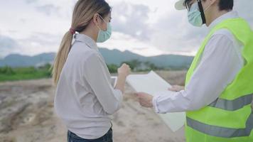 una donna proprietaria di terreni e manager ingegneri che discutono sui requisiti di costruzione in un paesaggio sterrato vuoto, assumendo un team di costruttori professionisti, tenendo un progetto di carta, possedendo un'attività di successo video