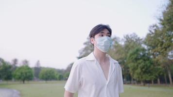 porträtt av ung asiatisk man i vit t-shirt utomhuspromenad i stadsparken, studentliv, generation z, glada fritidsmänniskor under pandemin, medicinsk coronavirus covid 19-mask, slow-motion-filmer video