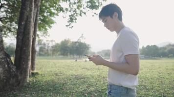 junger hellhäutiger Mann mit Smartphone beim Gehen im Park, Sonnenreflexion, heißes sonniges Tageslicht, moderner Lebensstil, SMS-Online-Dating senden, Chat-Messenger-Anwendung video