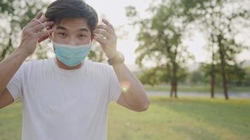 Closeup a young east asian man in gesturing of wearing protective face mask while standing inside of green public park, hands adjusting on mask ear loops, loose-fitting mask covers for nose and mouth video