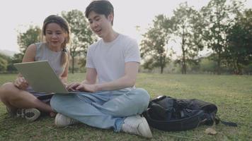 A young casual couple sitting on a green meadow inside public park, having a cheerful conversation on working project, hand using a smart technology, staring at computer, brainstorming giving an ideas video