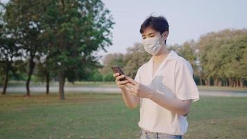 un hombre asiático de buen aspecto con una cara seria leyendo en un teléfono inteligente mientras camina en un parque verde, un hombre atractivo con un estilo de ropa mínimo, usando una máscara protectora cuando sale