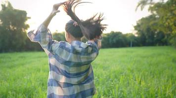 vista posteriore di una silhouette contadina sta fuori legando la sua lunga bruna contro la luce solare estiva e il campo di erba verde fresca, l'acconciatura femminile, la bellezza della natura e le persone, preparandosi per lavorare video