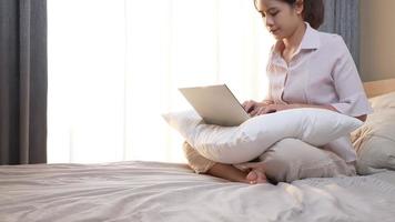 garota freelance atraente asiática em roupas casuais com um laptop no colo enquanto está sentado na cama confortável no quarto em casa com cortina branca contra a luz ensolarada, viajar e trabalhar com horário livre