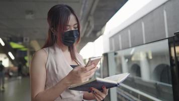 junge asiatische berufstätige frau trägt eine gesichtsmaske, die auf dem bahnsteig der u-bahn steht und mit dem handy nachrichten überprüft und die informationen sammelt, bevor sie zur arbeit geht, dame beschäftigt zu arbeiten, während sie auf den zug wartet video