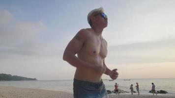 Strong muscular tan skin grey hair senior man wear sunglasses running on the beach during sun down, remote cardio exercise training on the island sea coast, outdoor exercises motivation, active senior video