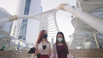 Asian business ladies walking together on the sky walk overpass showing city landscape after work, walking home with best friend, young two colleagues wear mask having conversation during a way home video