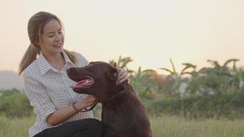 Young female sit down playing her friendly dark brown Labrador retrieve, pet care, walking dog at neighborhood street afternoon sunset light, bonding friendship family member, well trained dog hound video