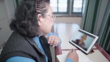 donna anziana asiatica indossare occhiali guardare tablet corso online lezione di classe, sedersi a scrivere con la matita sul taccuino, modello indossare maglione, generazione di anziani e tecnologia moderna stile di vita in pensione video