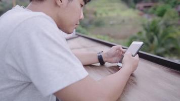 Der junge, moderne Mann genießt es, eine mobile App zu verwenden, sitzt auf einer langen Bank mit natürlichem Hintergrund, rutscht in sozialen Medien und schaut sich neue Feeds von Freunden an, während er sich entspannt, Technologie überall drahtlos video