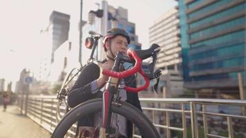 joven y feliz hombre de negocios asiático que lleva una bicicleta paseando por el puente de paso elevado de la ciudad a casa. luz del sol en el moderno edificio de cristal en el fondo, concepto de transporte y personas, ocio urbano video