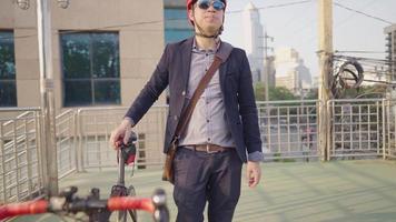 junger entspannender asiatischer mann, der sein fahrrad auf dem radweg schiebt, während er morgens zur arbeit geht, genoss das atmen der frischen morgenluft und die aktive aussicht, sonnenlichtsilhouette als hintergrund