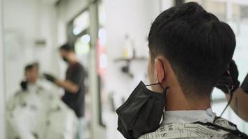 vista desde atrás dentro de la barbería de hombres, hombre asiático con máscara negra se corta el pelo. reflejo de espejo, patillas afiladas, caballeros peluqueros durante corona covid-19 reabrir negocios después de la pandemia video