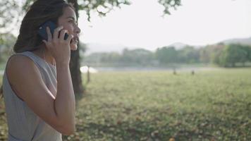 giovane donna di razza mista che parla sullo smartphone all'interno del parco estivo all'aperto, una tecnologia mobile wireless, l'uso delle tecnologie multimediali nelle relazioni a distanza, la società umana e l'ambiente video