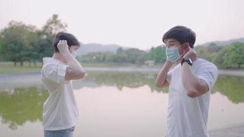 Two asian handsome man putting on facial mask while standing, next to pond park, against each other in the same time. Social distancing, Covid-19 prevention, surgical protective mask for corona virus video