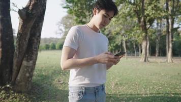 jovem homem asiático usando smartphone relaxando dentro do parque, parque ao ar livre ao ar livre, dia de sol quente, telefone de rolagem de cabeça para baixo, dispositivo portátil, telefone celular e vida diária moderna, faça logon no usuário do aplicativo video