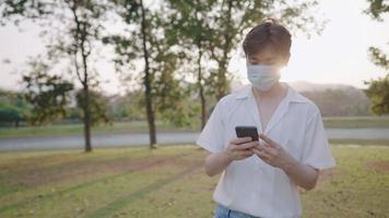 Young male wear face mask using smartphone standing alone inside the park at sunset, sun light flares reflection, modern lifestyle, send text message, online dating, application user log on member