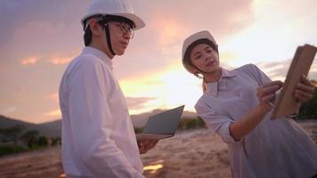 Asian surveying employee discussing on construction project at outdoor plain land site during sunset, working overtime, male and female colleagues, teamwork spirit brainstorming looking at tablet video