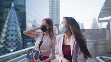 dos trabajadoras asiáticas vestidas formalmente en una nueva vida normal con máscara facial caminando al trabajo en la ciudad cruzando el puente con entornos urbanos circundantes, mujeres de negocios asiáticas, chicas hablan chismes video