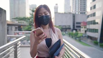 La joven y elegante mujer trabajadora asiática usa una mascarilla protectora usando un teléfono inteligente caminando por el sendero de la ciudad, la nueva vida normal se adapta a la nueva variante del virus, la hora pico ocupada, mostrando el área comercial de la vida