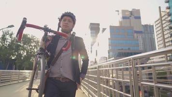 asiatischer selbstbewusster arbeiter trägt sein fahrrad auf der stadtüberführung, geschäfts- und umweltverkehrskonzept, sonnenreflexion auf modernem glasgebäude im hintergrund, stadtlandschaft video