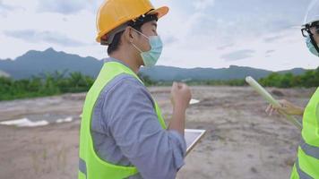 Two young engineering men wearing face protective mask while planning on working project at outdoor dirt construction site, self prevention during covid-19 pandemic, hiring professional builder team video