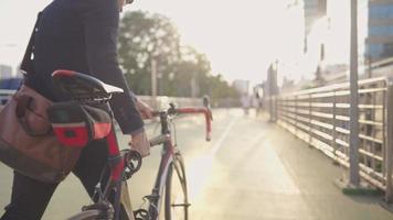 joven trabajador asiático inteligente vestido formalmente caminando al aire libre y llevando bicicleta hacia adelante, paisaje urbano, puesta de sol en frente como fondo, ciclista masculino paseando y sosteniendo bicicleta, pasatiempo y estilo de vida video