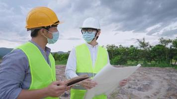 asiatische architektur- und ingenieurarbeiter tragen sicherheitsweste und schutzhelm, schützende gesichtsmaske, die während einer pandemie auf der baustelle zusammenarbeiten, tablet-blaupausenpapier halten, soziale distanz video
