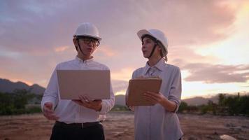 asian construction worker discussing on building construction project at outdoor plain land site during sunset, working overtime, Real Estate investing innovation project, male and female architecture
