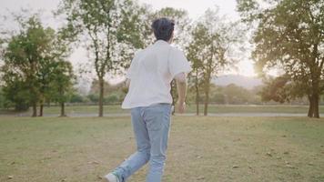 Rear view of young asian man slowly running to sunlight behind trees, man runs on a wide meadow field inside the open air park, male casual clothing, wearing denim pant, happy people and lifestyle video