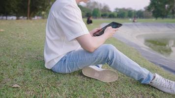 Young male freelance worker sit down on comfortable grass inside public park during day, hands holding digital tablet with using fingers on touch screen, mobile technology, peace and relaxation place
