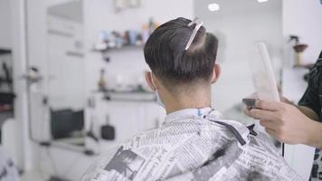 homem asiático de visão traseira corta o cabelo durante o bloqueio de quarentena, barbearia profissional, pequeno empreendedor de negócios de inicialização, barbeiro masculino reabrindo negócios, cabeleireiro aparando usando máquina de cortar cabelo