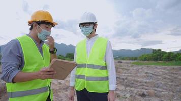 Aziatische bouwvakkers dragen een medisch gezichtsmasker en een volledig veiligheidsuniform dat samenwerkt tijdens pandemie op de bouwplaats buiten, tablet vasthoudt met rapportage aan manager, milieuvriendelijk project video