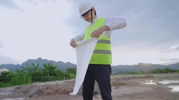 Asian male Engineer architecture wear safety vest helmet folding blueprint drawing paper after finishing survey work, specialist expertise working field, over time hard working employee, outdoor site video