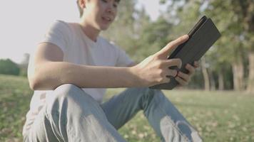 A young happy casual man siting knees up on a green meadow enjoying having text conversation with friend inside a natural park, hot summer air, laughing with tablet on hands, outdoor leisure activity video