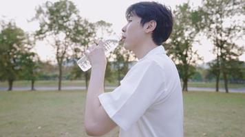 joven y apuesto hombre asiático bebiendo agua mientras se relaja en el parque al aire libre con puesta de sol a distancia, sintiéndose fresco con el concepto de humedad saludable de la piel, rehidratación agua mineral vitaminada, vista lateral