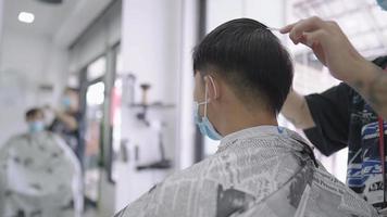 un hombre asiático usa una mascarilla protectora para cortarse el cabello durante la pandemia de cuarentena, una cuchilla eléctrica para cortar el cabello, un peluquero masculino que reabre el negocio, creación de un diseñador de cabello masculino, trabajo de servicio al cliente video