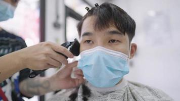 junger asiatischer junge trägt gesichtsmaske, bekommt haare geschnitten, bleibt geschützt und sicher außerhalb des hauses, männliche friseurläden kämpfen, kleine unternehmen werden wiedereröffnet, kerl haardesigner, kundenhygiene fühlen sich sicher video