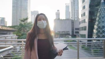 portrait heureux femme d'affaires asiatique confiante marchant et souriant à la caméra, contre un bâtiment en verre moderne et d'autres architecture derrière, belle employée de bureau portant un masque médical à l'extérieur