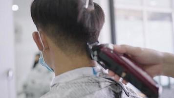 barbeiro asiático corta cabelo com aparador de cabelo elétrico, vista traseira da cabeça do cliente jovem na reabertura da barbearia masculina de negócios, serviço de corte de cabeleireiro, use máscara facial para prevenção de doenças infecciosas video