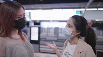 Two young adult asian women wear protective facemask talking to each other on the skytrain platform while que up waiting for the train, new normal life after pandemic, people adapt to new lifestyle