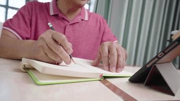 Close up mature male adult working from home with tablet, writing with pencil on the notebook, wireless technology distancing remote workplace, stay at home quarantine, old age man and modern life video