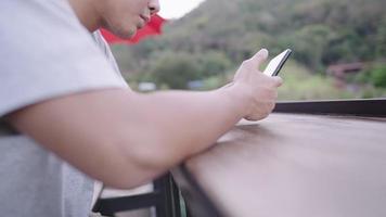 Half face shooting, Young male playing on smartphone while relaxing with green natural in background, asian white man turns face and talking to someone next to him on long bench before accept call