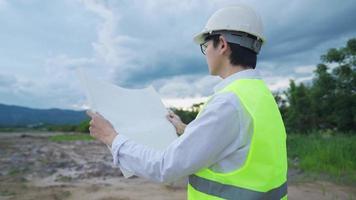 Young professional senior worker reading a building structure on blueprint while standing on rural rough land, hands holding equipment development plan, safety wearing worker, construction worker wear